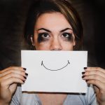 Woman with tear-stained eye makeup holds up a sign with a hand-drawn smile to her mouth